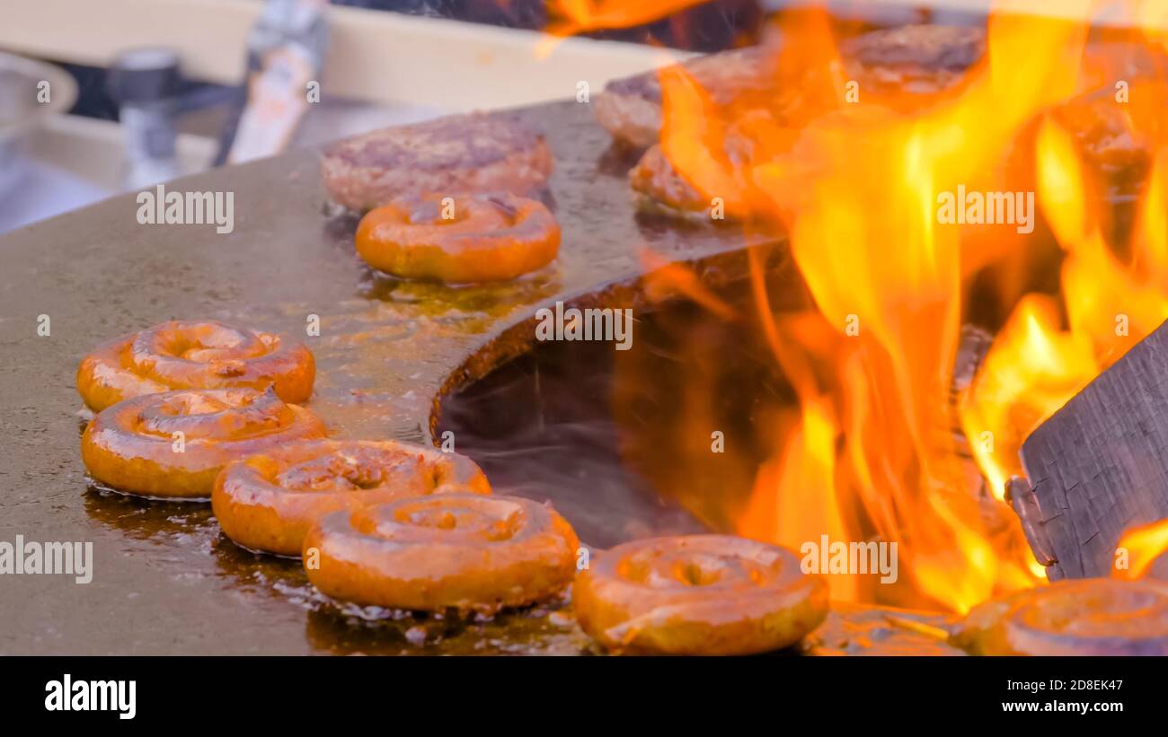 Prozess des Grillens von frischen Spiral Schweinefleisch Würstchen auf Brazier mit heißer Flamme auf Sommer lokalen Lebensmittelmarkt - Nahaufnahme. Kochen im Freien, Grill Stockfoto