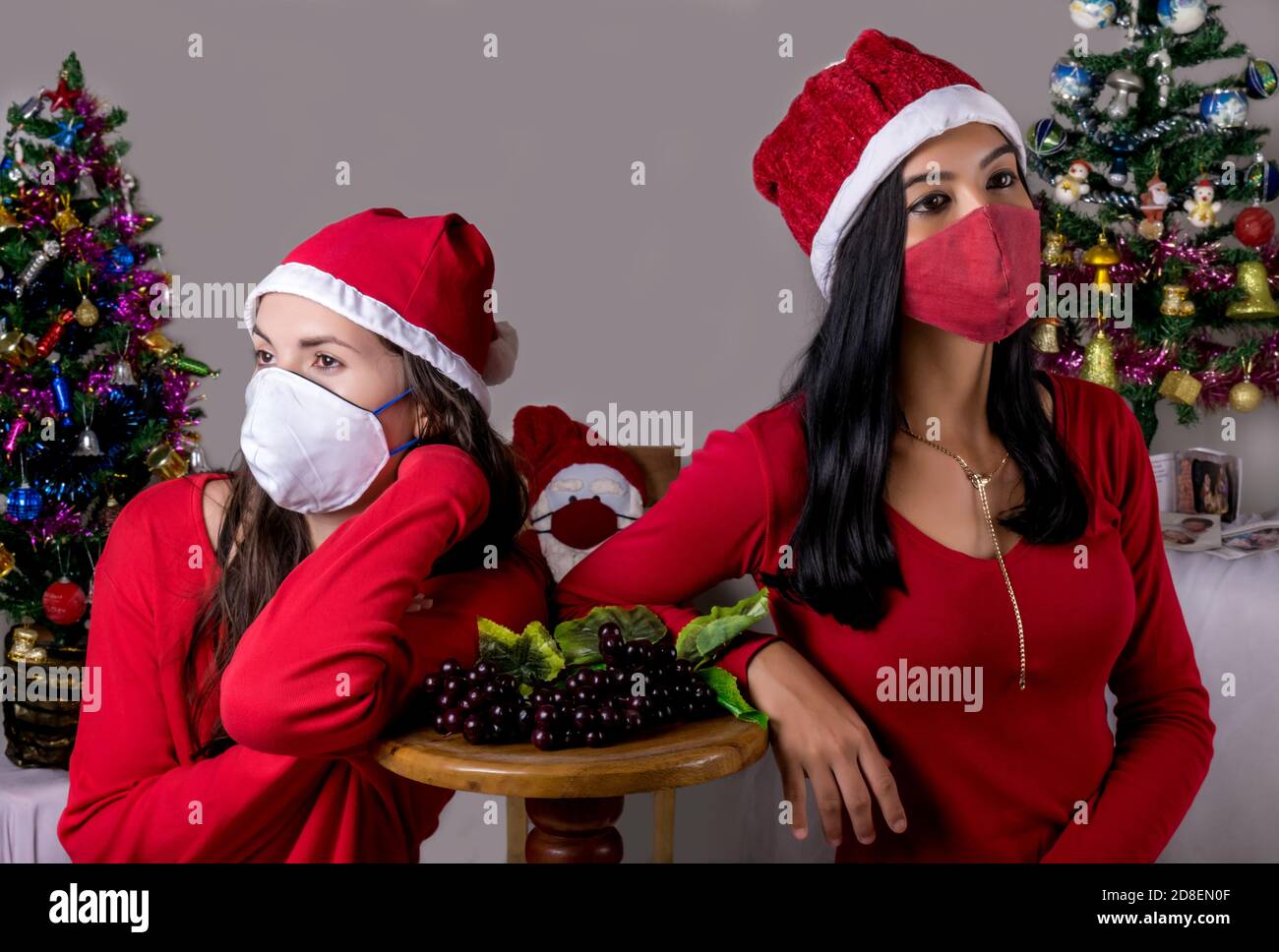 Zwei Mädchen in weihnachtlicher Umgebung, die Masken tragen, die sich jeweils dem anderen zuwenden und ihre Arme auf einen kleinen Holztisch und einen Weihnachtsmann in der BA lehnen Stockfoto