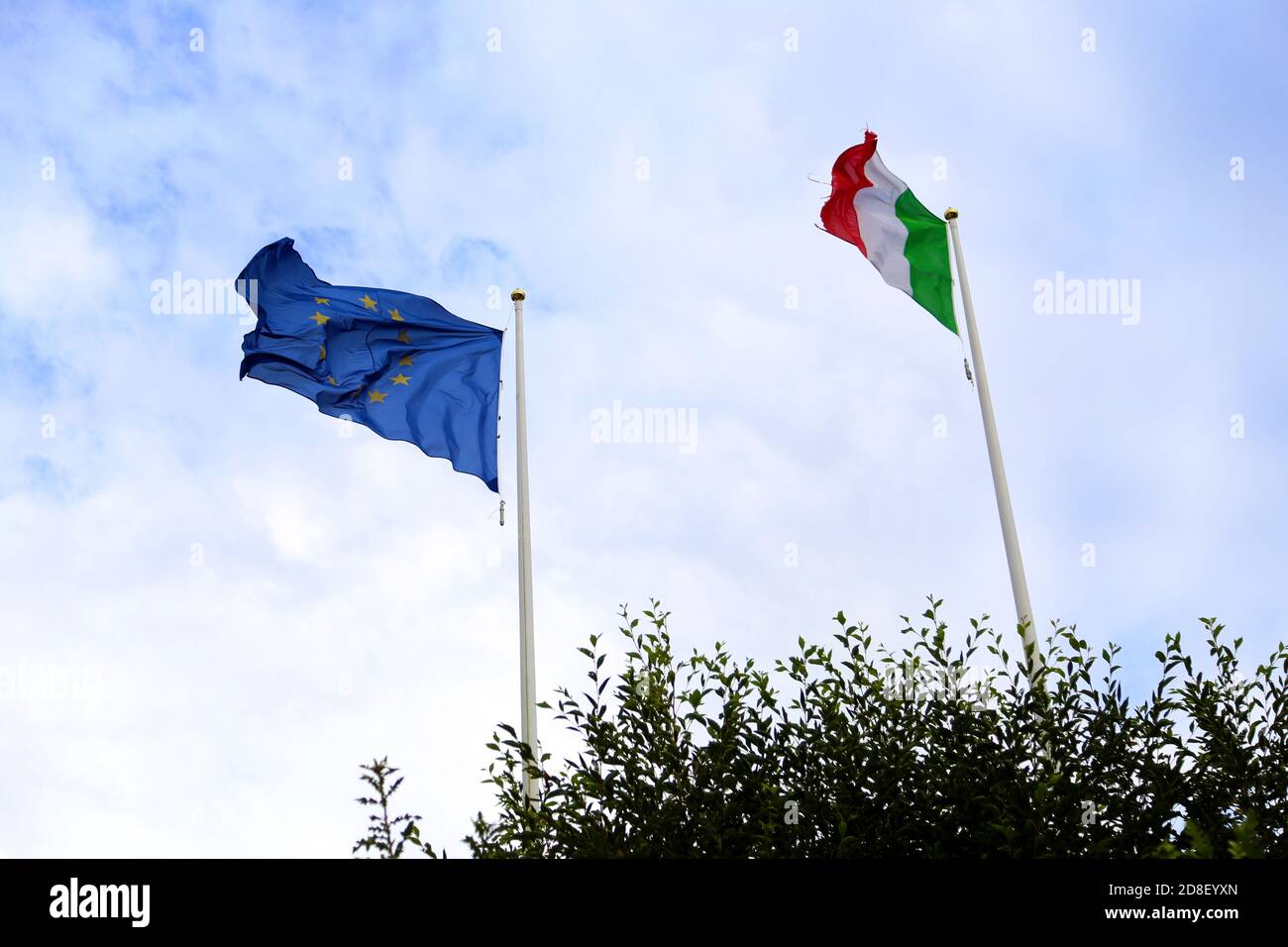 Die Flaggen Europas und Italiens wehen gegen einen teilweise bewölkten Himmel. Die beiden Fahnenmasten stehen hinter der Vegetation Stockfoto