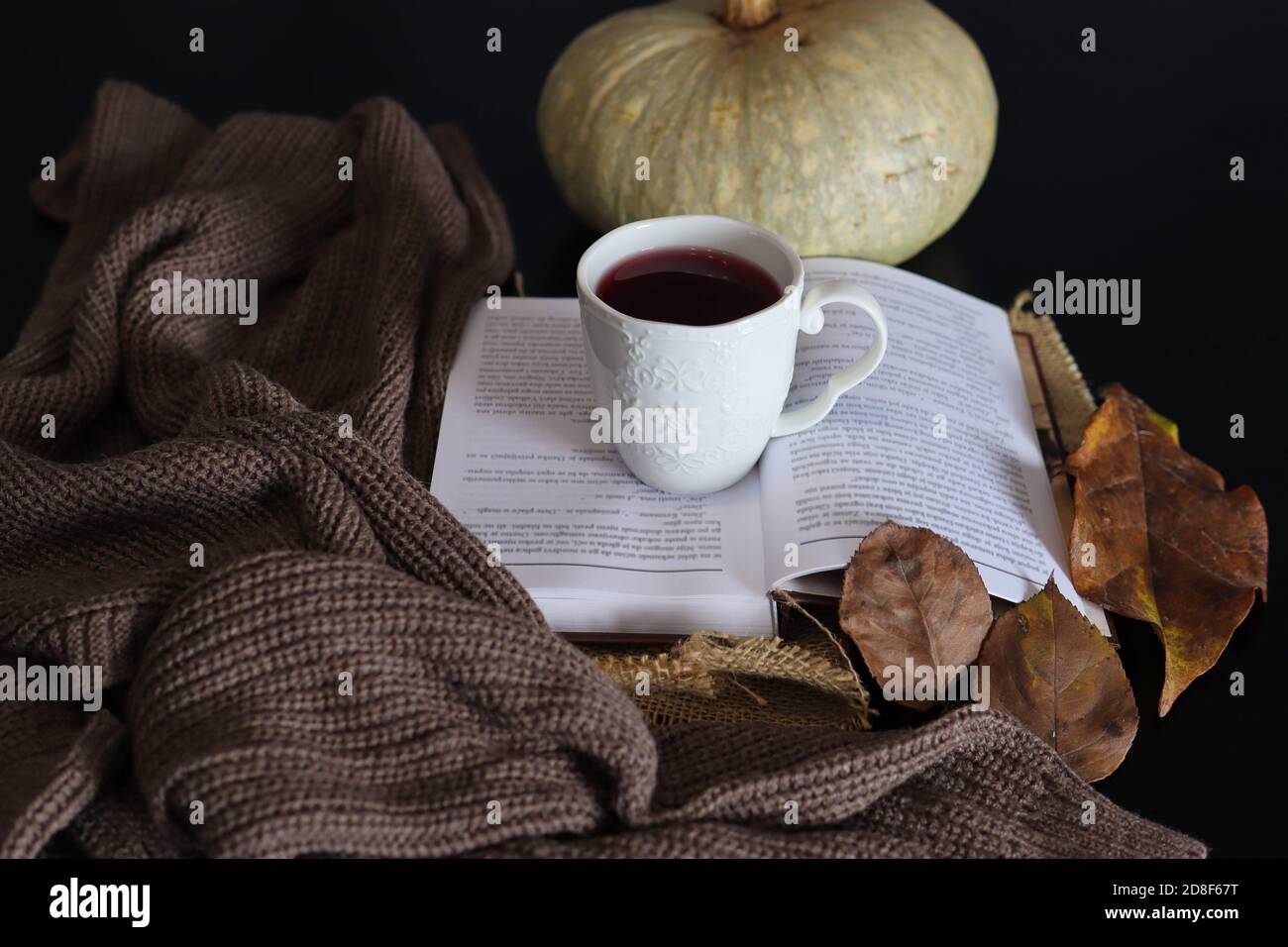 Eine Tasse heißen Tee auf einem offenen Buch und einem dekorierten Schreibtisch mit Schal und trockenen Blättern. Heißgetränk und Herbstsaison Konzept Hintergrund Stockfoto