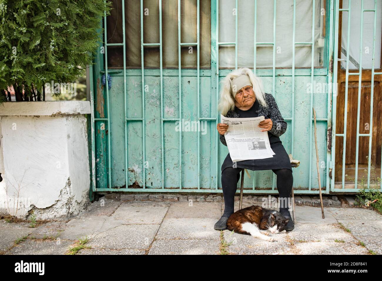 Eine ältere Frau liest die Zeitung vor ihrer Haustür, während ihre Katze zu ihren Füßen in Mzcheta, Georgien, Kaukasus, Osteuropa schläft. Stockfoto