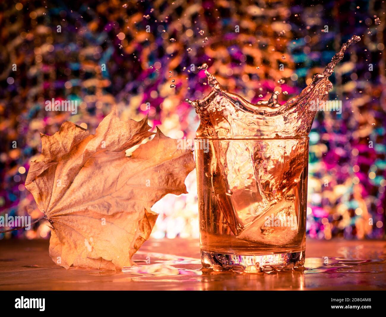 Eiswürfel spritzen in Whiskey-Glas mit Ahornblatt Farbenfroher Hintergrund Stockfoto