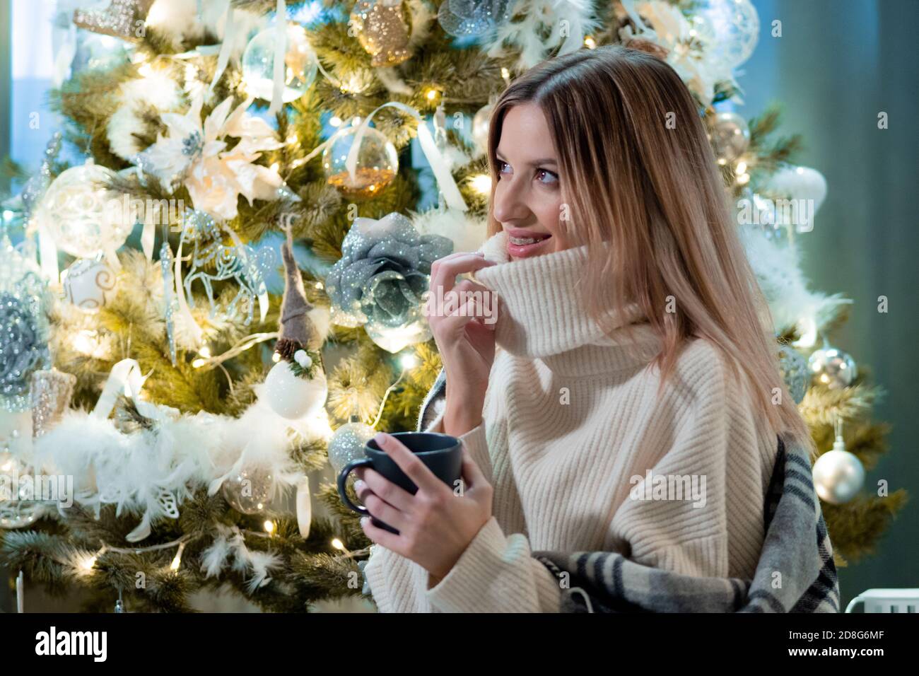 Neues Jahr zu Hause. Eine junge Frau in einem gemütlichen weißen Pullover sitzt auf dem Boden neben einem funkelnden Weihnachtsbaum und trinkt ein heißes Getränk Stockfoto