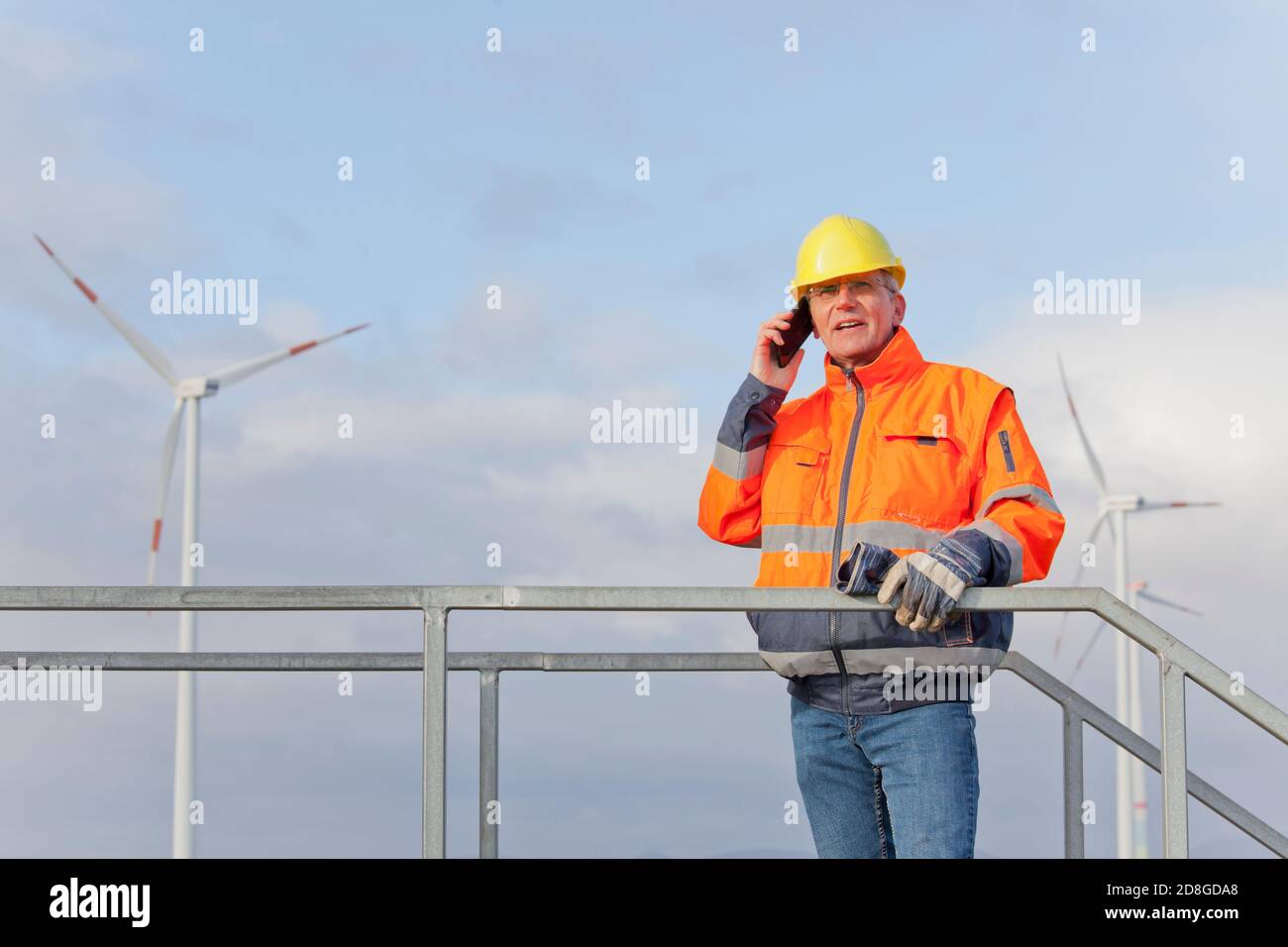 Techniker mit Schutzarbeiten tragen Gespräch auf Mobiltelefon in Vorderseite der Windenergieanlagen im Hintergrund Stockfoto
