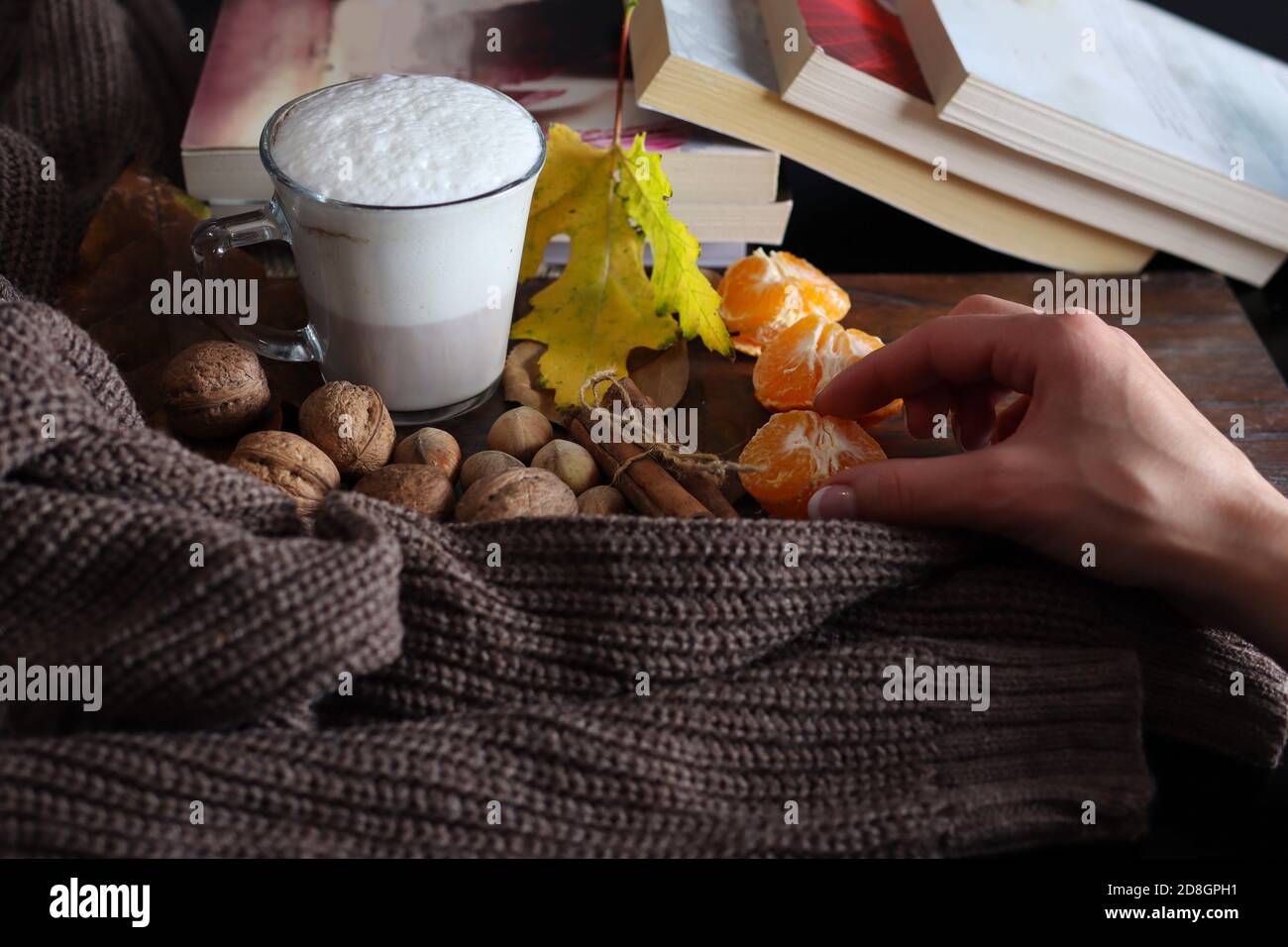 Die Mädchenhand nimmt Orangenfrüchte vom dekorierten Schreibtisch mit einer Kaffeetasse, Haselnuss, Walnuss und Pullover. Herbst und gesundes Lebensmittelkonzept Stockfoto