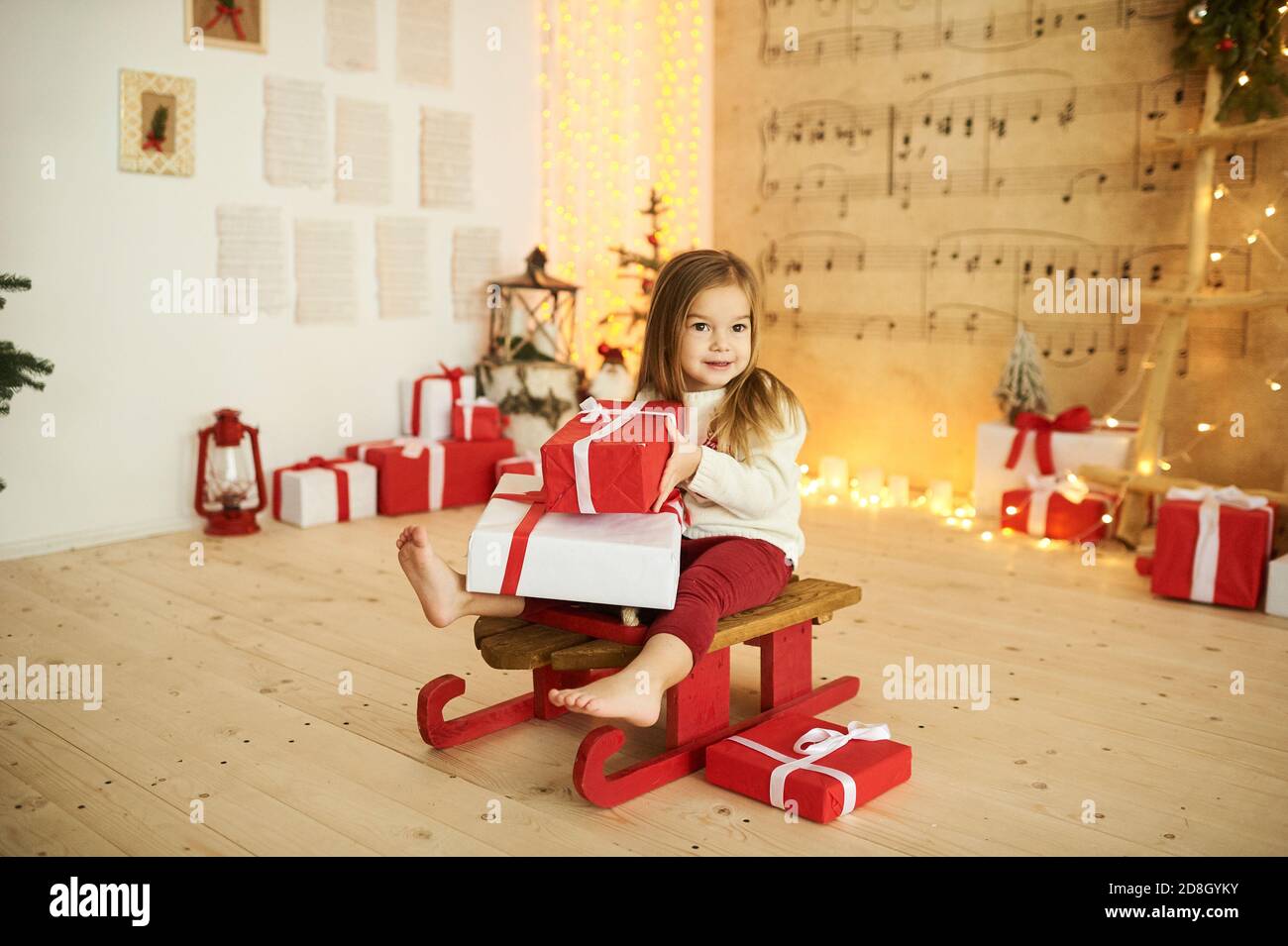 Ein Porträt eines kleinen Mädchens auf einem roten Schlitten Mit einem Geschenk auf einem verschwommenen Licht Hintergrund Stockfoto