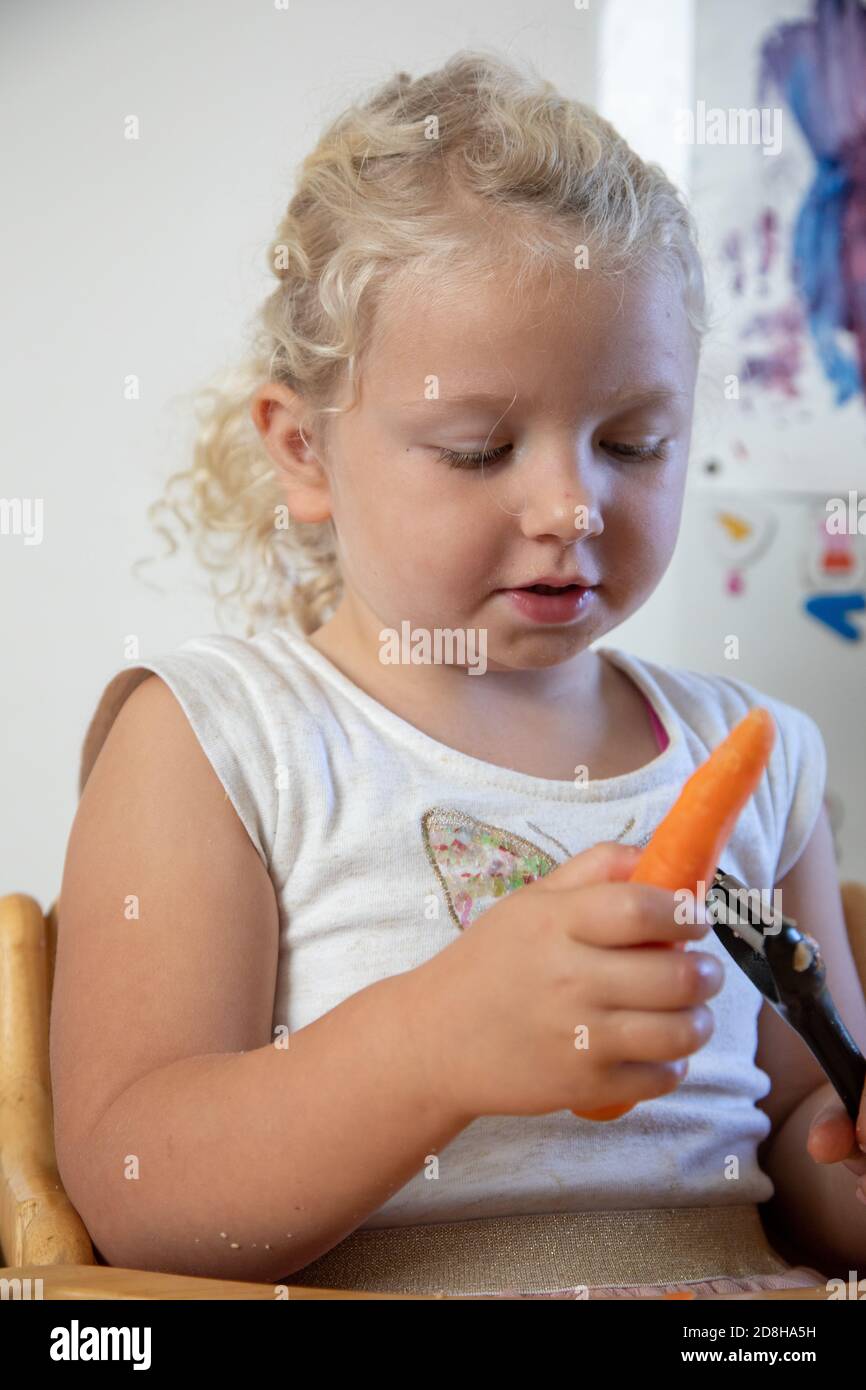 Ein dreijähriges Mädchen schält eine Karotte selbstständig in sich Wohnküche Stockfoto