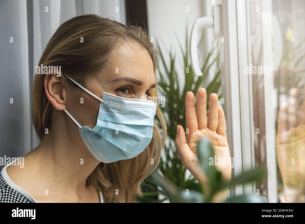 Home Quarantäne Virus Pandemie - junge Frau mit medizinischen Gesicht Maske schaut durch das Fenster und winkt mit der Hand zum Freund Stockfoto