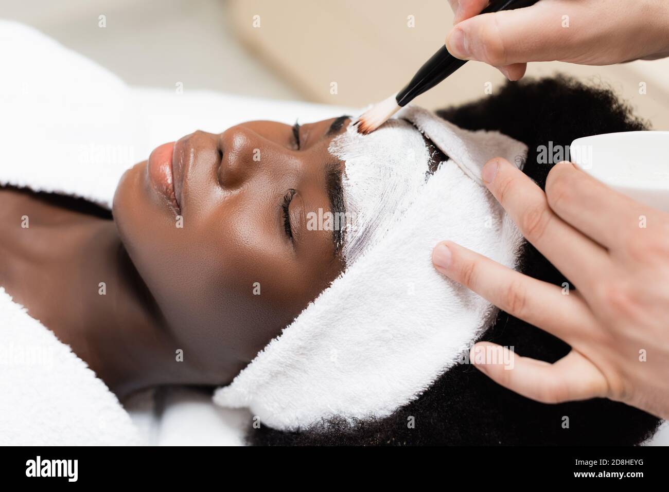 Nahaufnahme des Mannes, der Gesichtsmaske mit Kosmetik anwendet Pinsel auf der Stirn der afroamerikanischen Frau im Spa-Salon Stockfoto