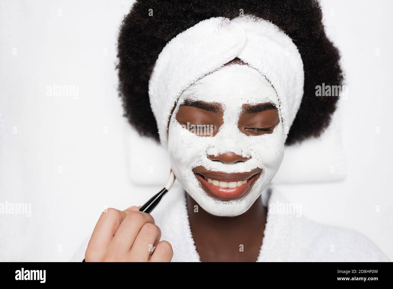 Draufsicht auf lächelnde afroamerikanische Frau mit Gesichtsmaske In der Nähe von Spa-Therapeuten mit Kosmetikbürste im Spa-Salon Stockfoto
