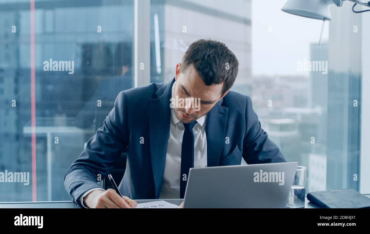 Erfolgreicher Geschäftsmann sitzt an seinem Schreibtisch, arbeitet mit Papieren, unterzeichnet Dokumente und mit Laptop. Im Fenster Big City Business District View. Stockfoto