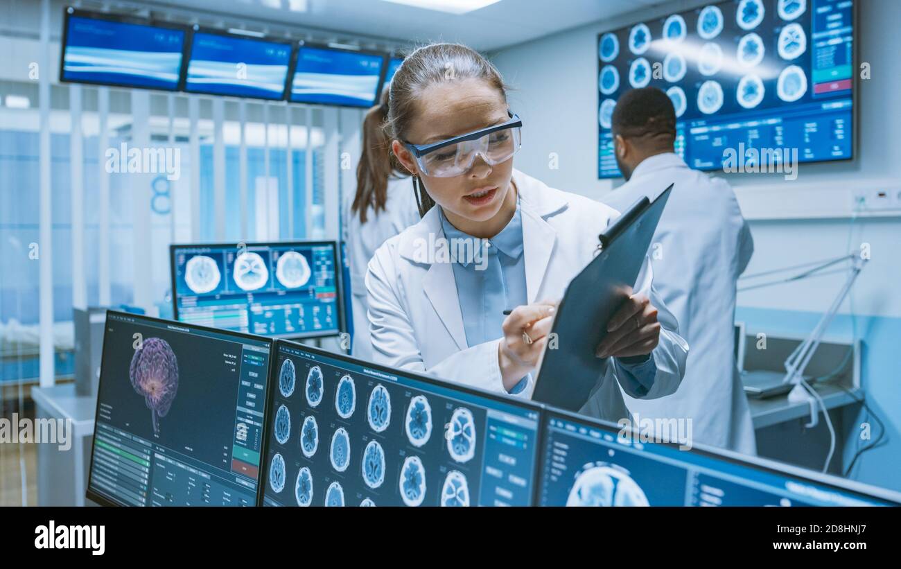 Senior Medical Scientist berät weibliche Lehrling mit Clipboard, Er arbeitet mit CT Brain Scan Bilder auf einem Personal Computer im Labor Stockfoto