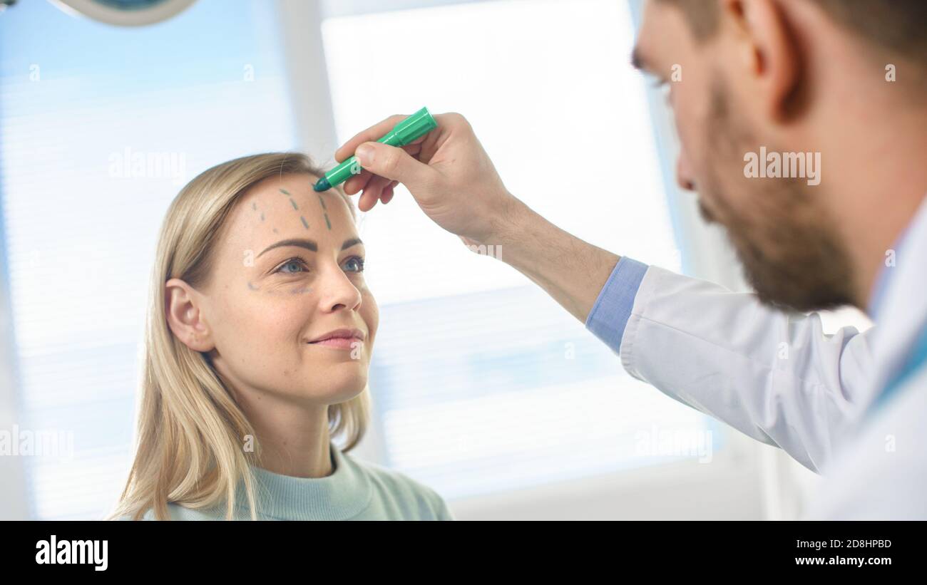 Over the Shoulder Shot eines plastischen Kosmetikchirurgen zeichnet Linien auf ein Gesicht eines schönen weiblichen Modells für die Facelift-Operation. Stockfoto