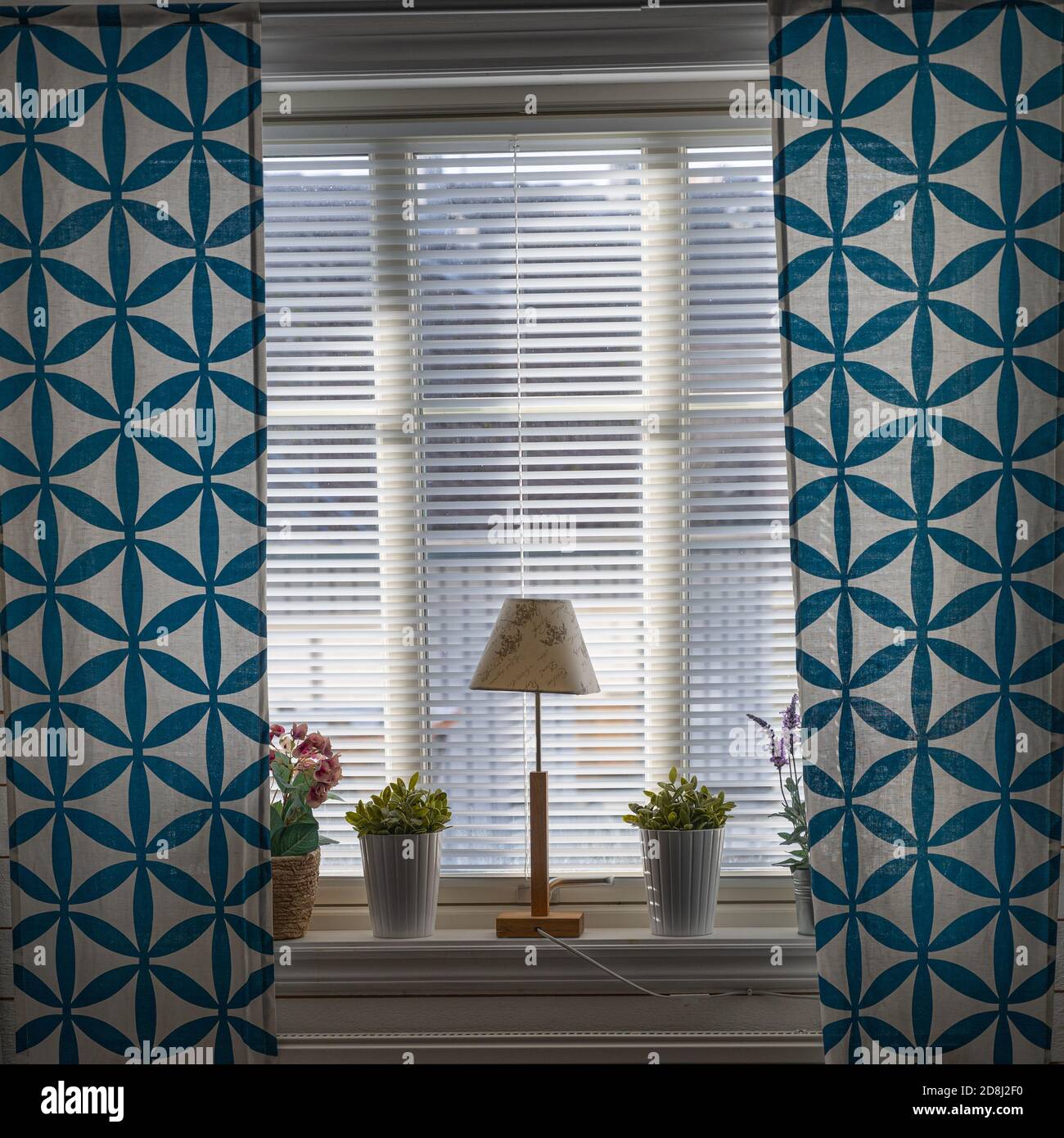 Ein Fenster mit blauen Vorhängen, einer Lampe und Blumen. Schönes Licht kommt durch die Jalousien Stockfoto