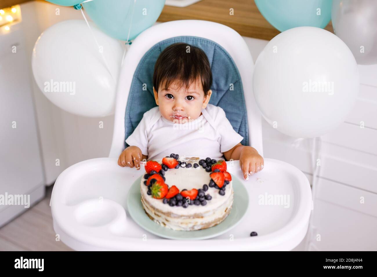 Kleiner Junge, der in einem Hochstuhl in der weißen Küche sitzt Und Verkostung ersten Jahr Kuchen mit Früchten auf Hintergrund mit Ballons Stockfoto