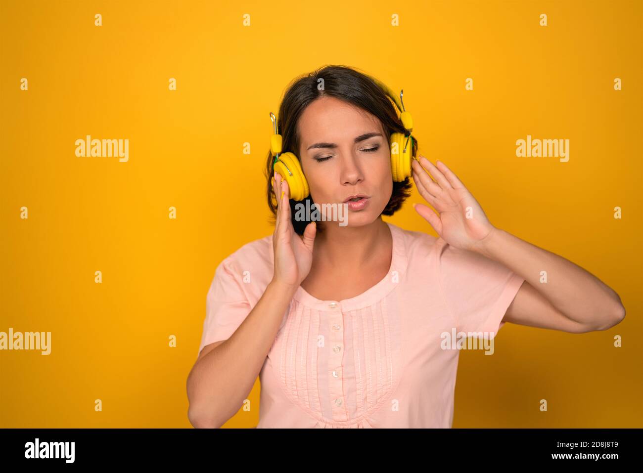 Glückliche Frau hört Musik in gelben Ohrenklappen. Kaukasische weibliche Modell trägt rosa T-Shirt isoliert auf gelbem Hintergrund. Stockfoto