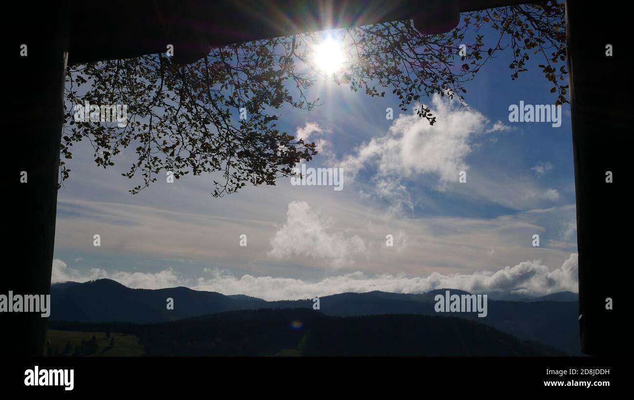 Blick durch ein Fenster von einem Holzhaus in der Wald mit der Sonne am Himmel umgeben von Wolken Und mit dem Schwarzwald im Hintergrund Stockfoto