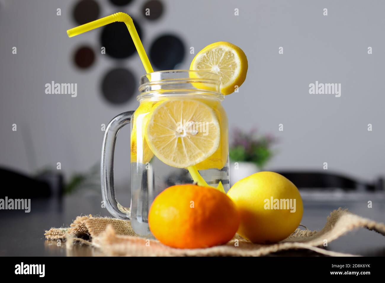 Limonadengetränk in einem Glasgefäß mit Zitronenscheiben und einem Trinkschlauch auf einem dekorierten Holzschreibtisch mit Zitronen- und Orangenfrüchten. Gesunde Getränke Konz Stockfoto