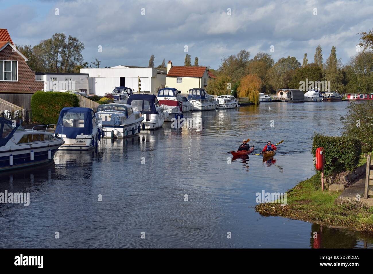 Zwei Kajakfahrer passieren festgemachtes Vergnügungsboote und Yachten entlang des Flusses Waveney in der Stadt Beccles in Suffolk, England. Teil der Suffolk Broads. Stockfoto