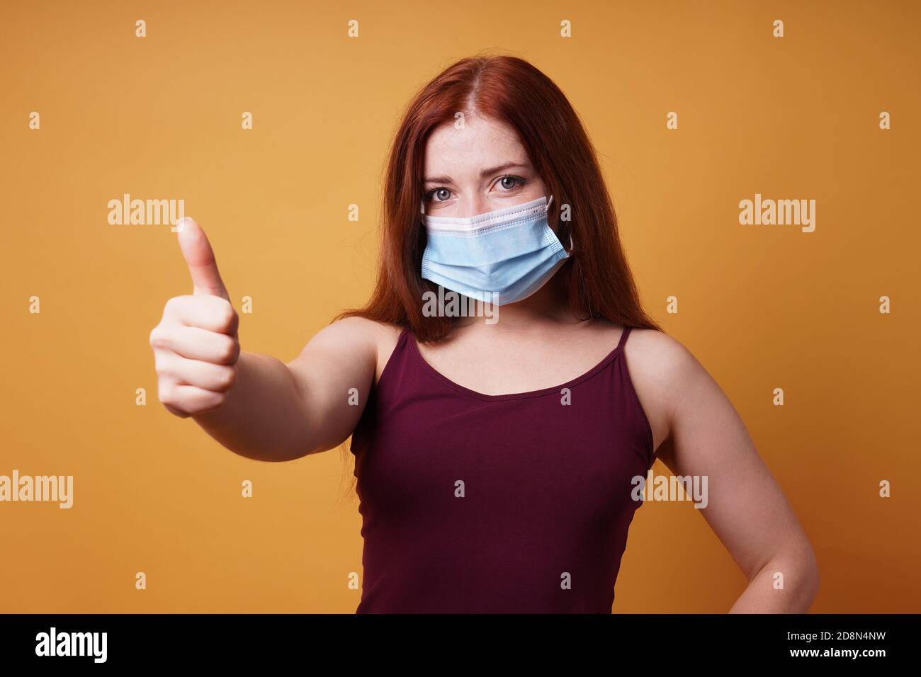 Junge Frau trägt eine medizinische Gesichtsmaske richtig bedeckt Mund Und Nase und geben Daumen hoch - Schutz gegen Corona Virus - Studio Portrait auf orangenen Hintergrund mit Kopie Platz Stockfoto