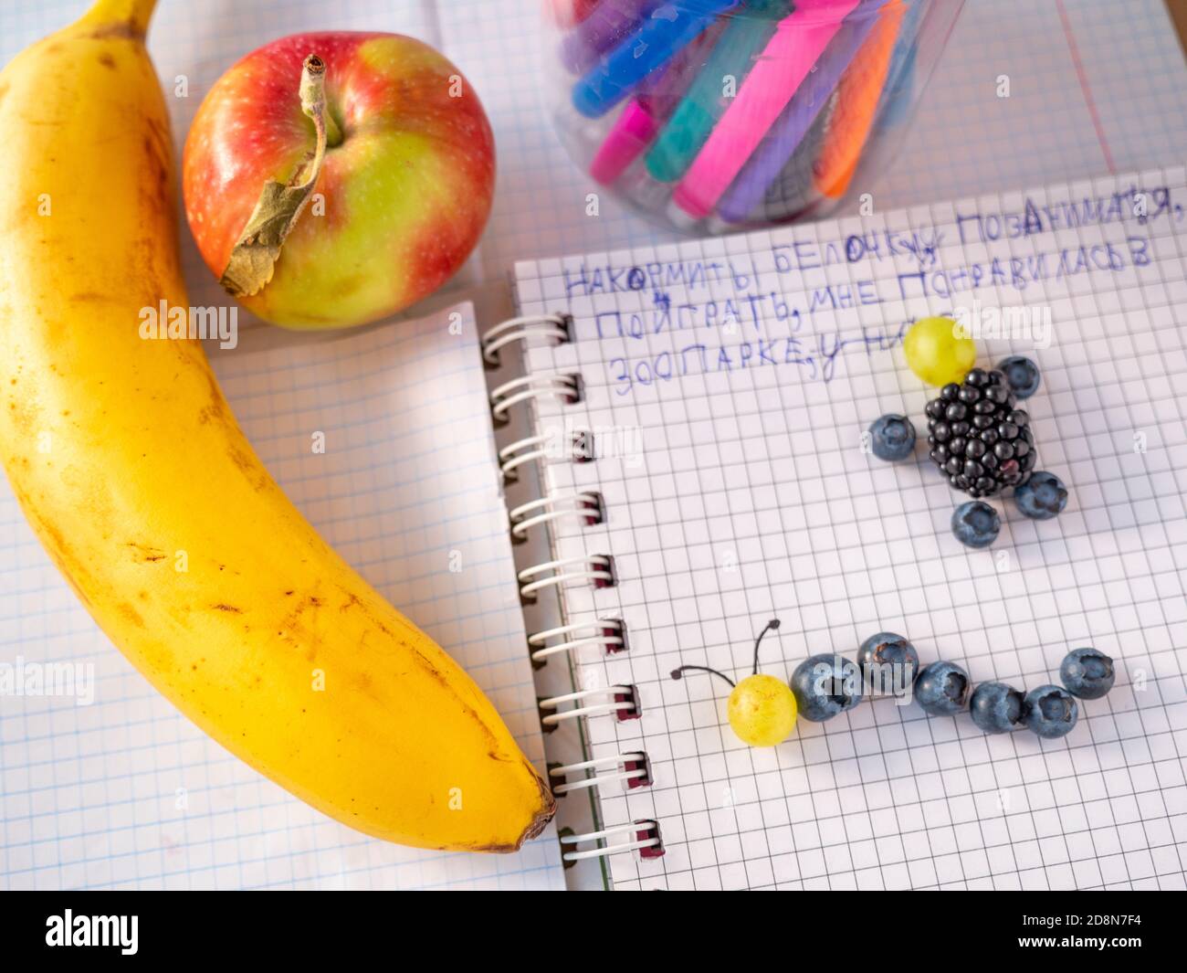 Fröhliche Kinder lernen ohne langweilig beim Spielen leckere Spiele. Handwerk und gesund essen. Apfel-, Banane- und Buntstifte und Stifte in einem Plastikglas. Stockfoto