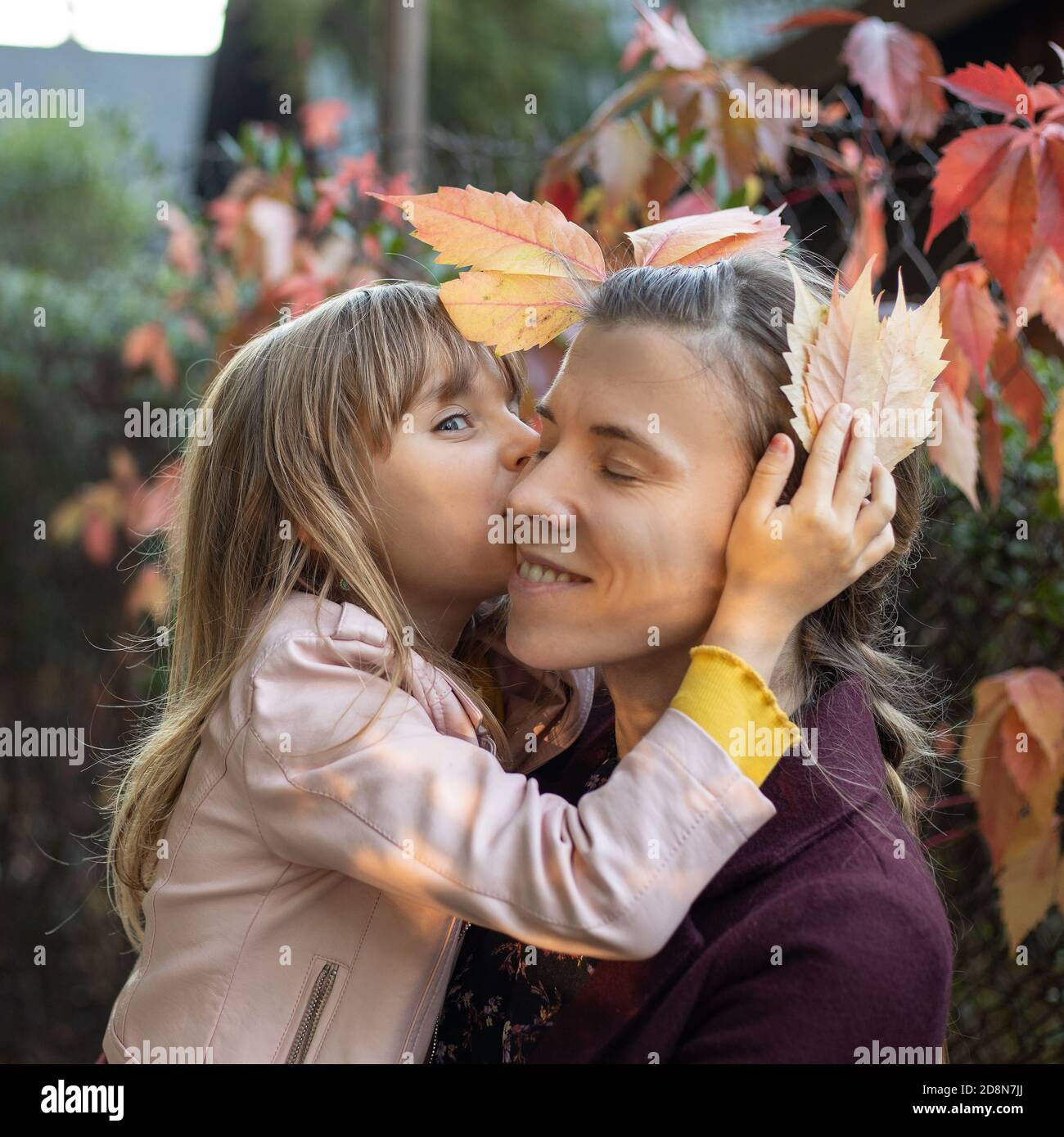 Ein kleines Mädchen von 7 Jahren küsst und umarmt ihre Mutter vor dem Hintergrund der Herbstblätter im Freien. Das Konzept der glücklichen Mutterschaft Stockfoto