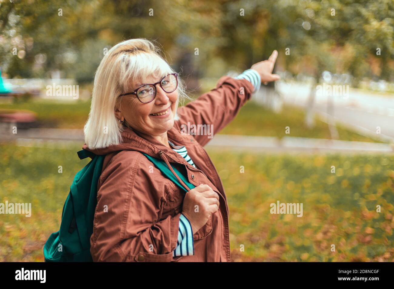 Erwachsene blonde Frau in bequemer Kleidung mit Rucksack zeigt die Richtung im Park mit dem Finger. Touristenreisender in einem wunderschönen Ort Stockfoto