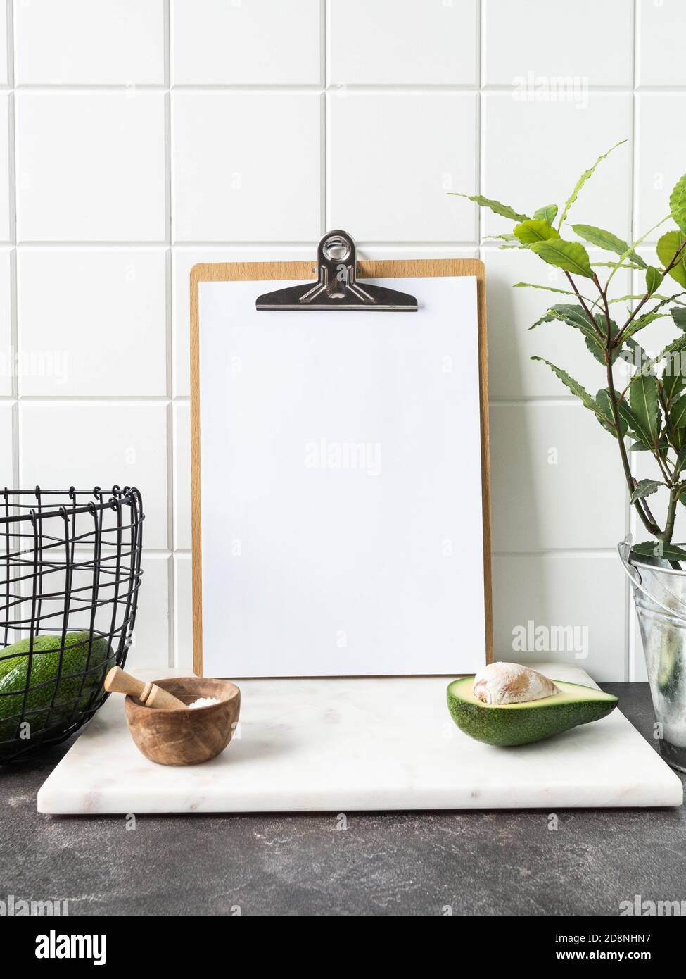 Stilvoller weißer Küchenhintergrund mit Korb mit Avocado, Lorbeer-Pflanze in Topf und Zwischenablage mit leerem Blatt Papier für Text, Kopierraum, Front vie Stockfoto