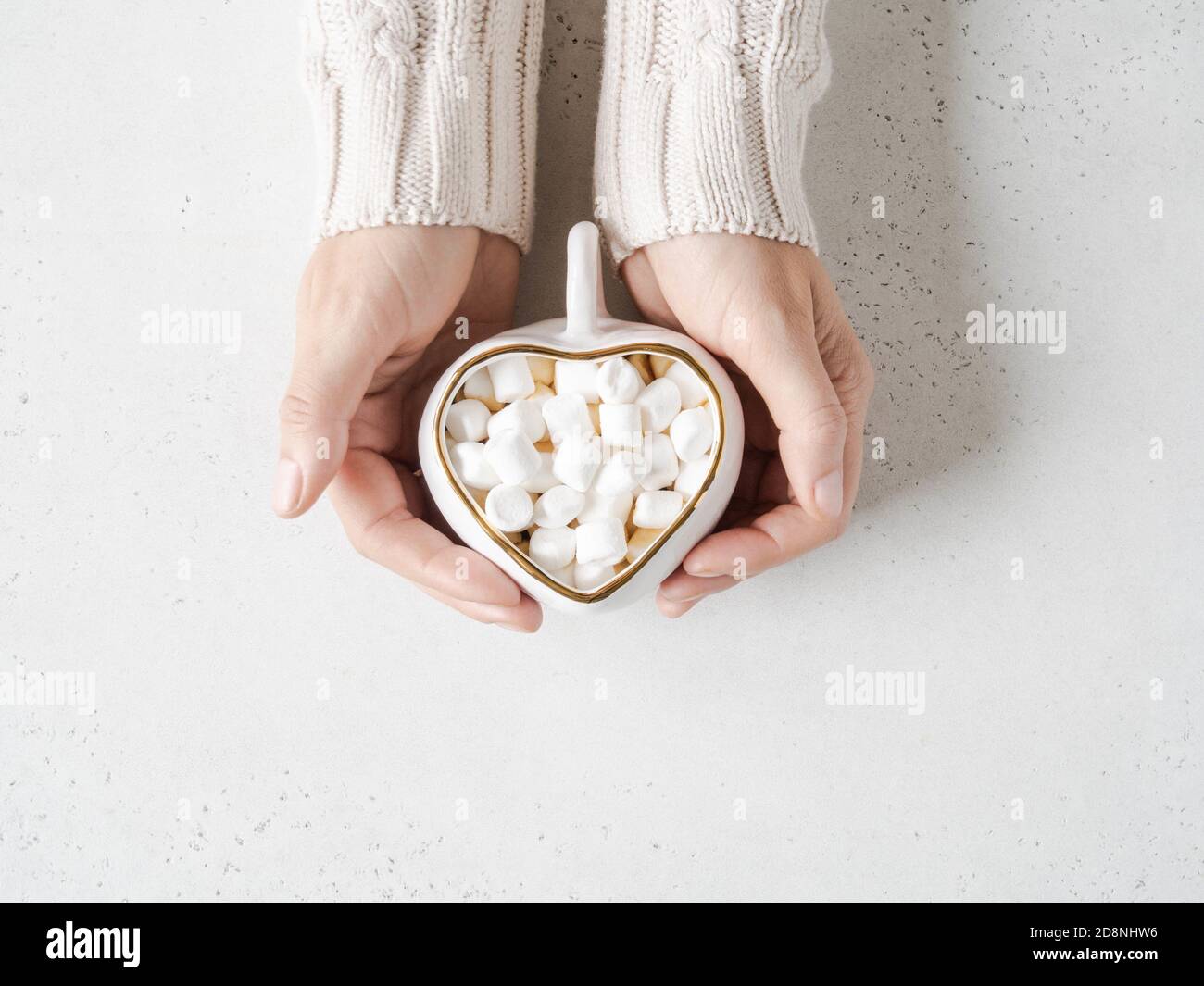 Weibliche Hände halten weiße Keramik-Tasse in Form von Herz mit heißem Getränk und Marshmallows auf hellem Hintergrund. Draufsicht. Speicherplatz kopieren Stockfoto