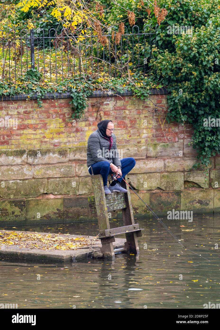 Ein Mann, der auf einem hölzernen Pfosten neben einem Fluss sitzt und einen Kapuzenpullover trägt, der auf einem Tor sitzt und mit einer Angelrute angeln kann. Stockfoto