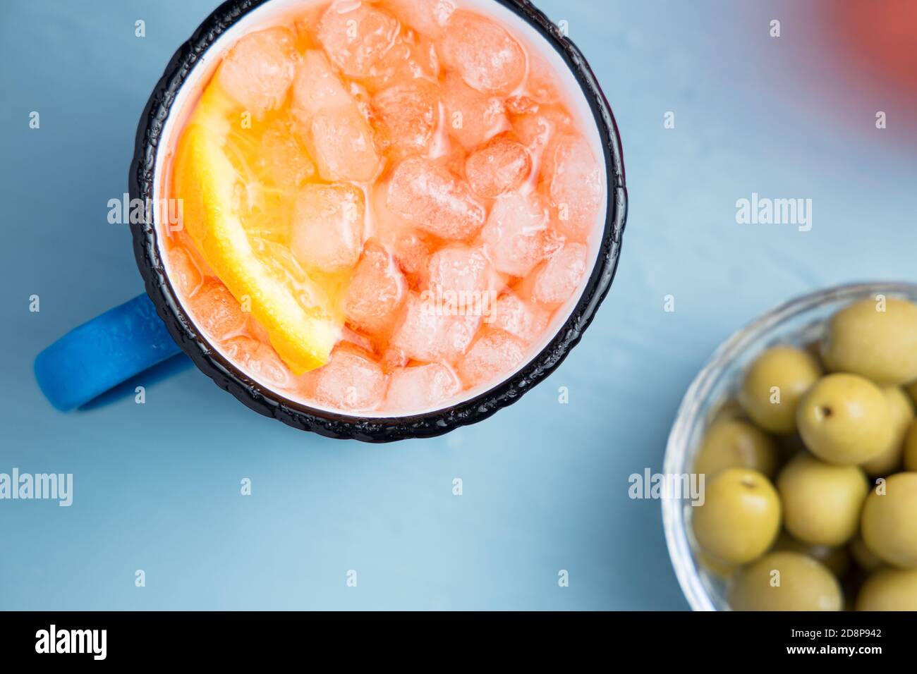 Emaillierter Becher des klassischen italienischen Aperitifs Spritz Cocktail mit Scheibe Orange und Oliven auf einem blauen Tisch. Draufsicht Stockfoto