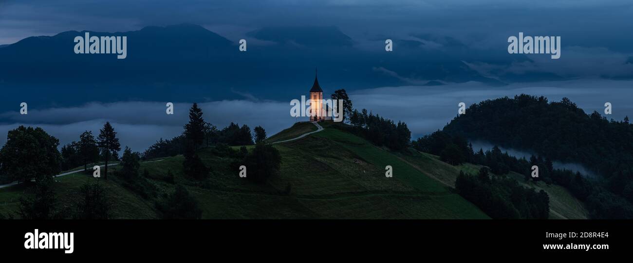 Jamnik, Slowenien - Panoramablick auf die blaue Stunde in Jamnik mit beleuchteten St. Primoz-Kirche auf einem Hügel bei einem nebligen Sommeranbruch. Die Julischen Alpen im Hintergrund Stockfoto