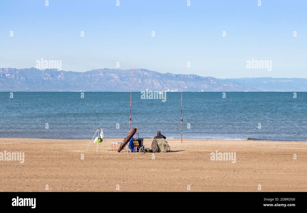 Mann Angeln am Strand mit zwei Ruten und andere Angelausrüstung, ist landbasierte Fischerei ein entspannendes Hobby. Berge und Boote im Hintergrund. Delta de Stockfoto