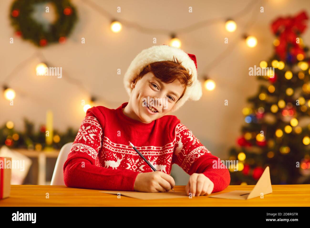 Lächelnder Junge in rotem Hut und Pullover sitzen und schreiben Brief an den Weihnachtsmann mit Wünschen und Träumen Stockfoto