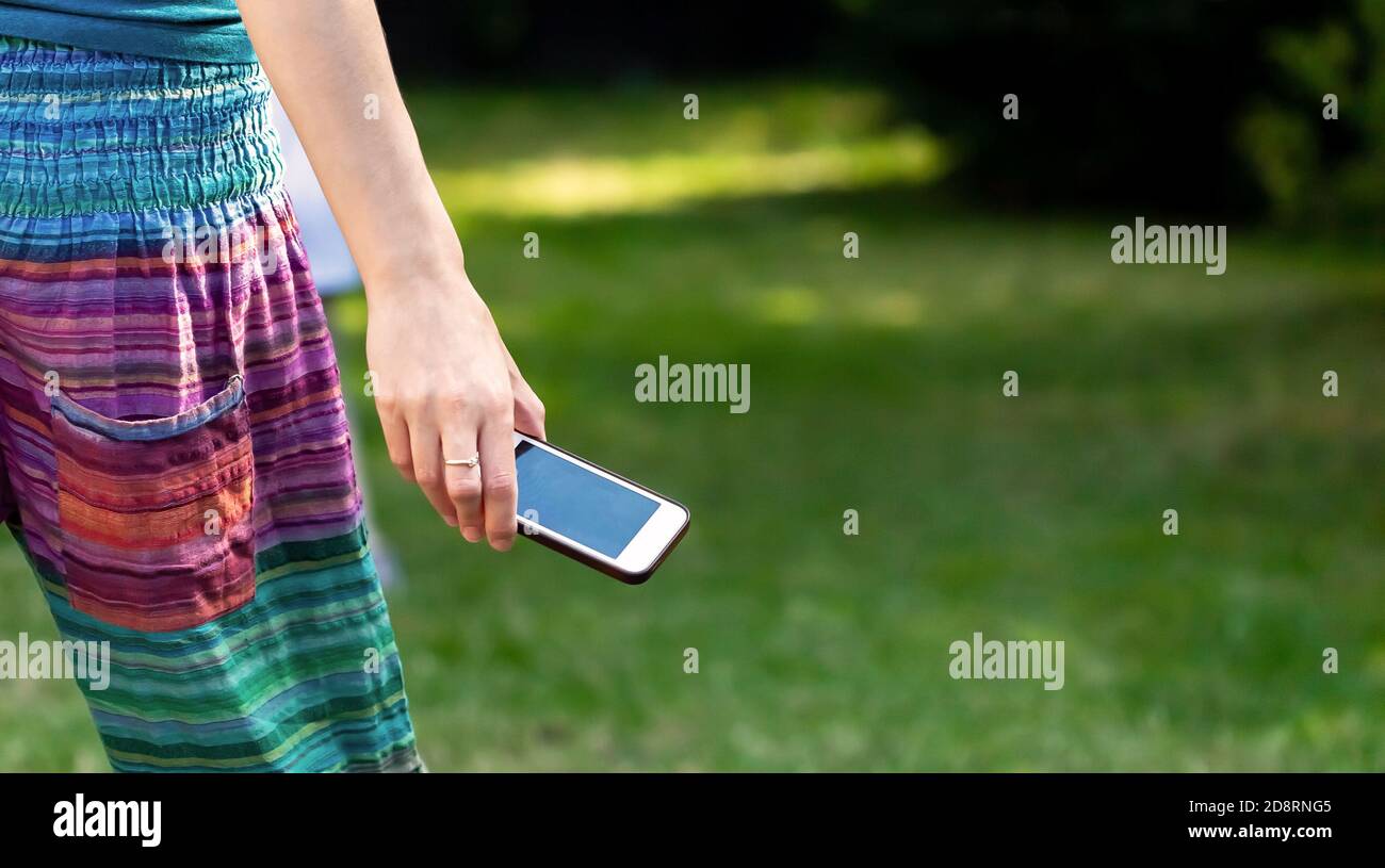 Frau hält ein Smartphone in der Hand, im Freien, grüner Garten im Hintergrund, Weitwinkel, Kopierraum. Ring am Finger. Junge anonyme Mädchen Stockfoto