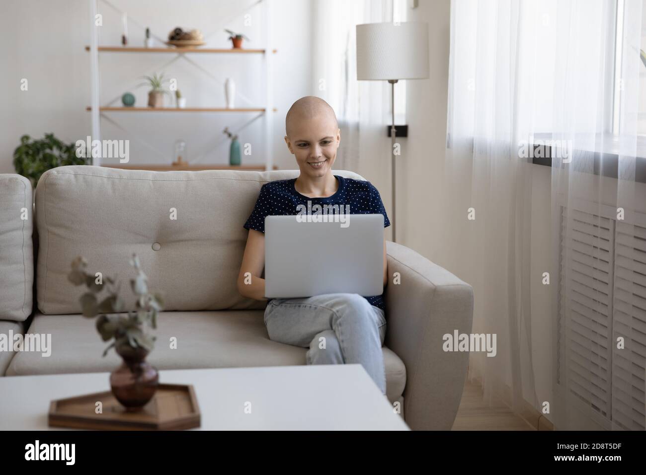 Lächelnde haarlose Frau mit Laptop, sitzt auf gemütliche Couch Stockfoto