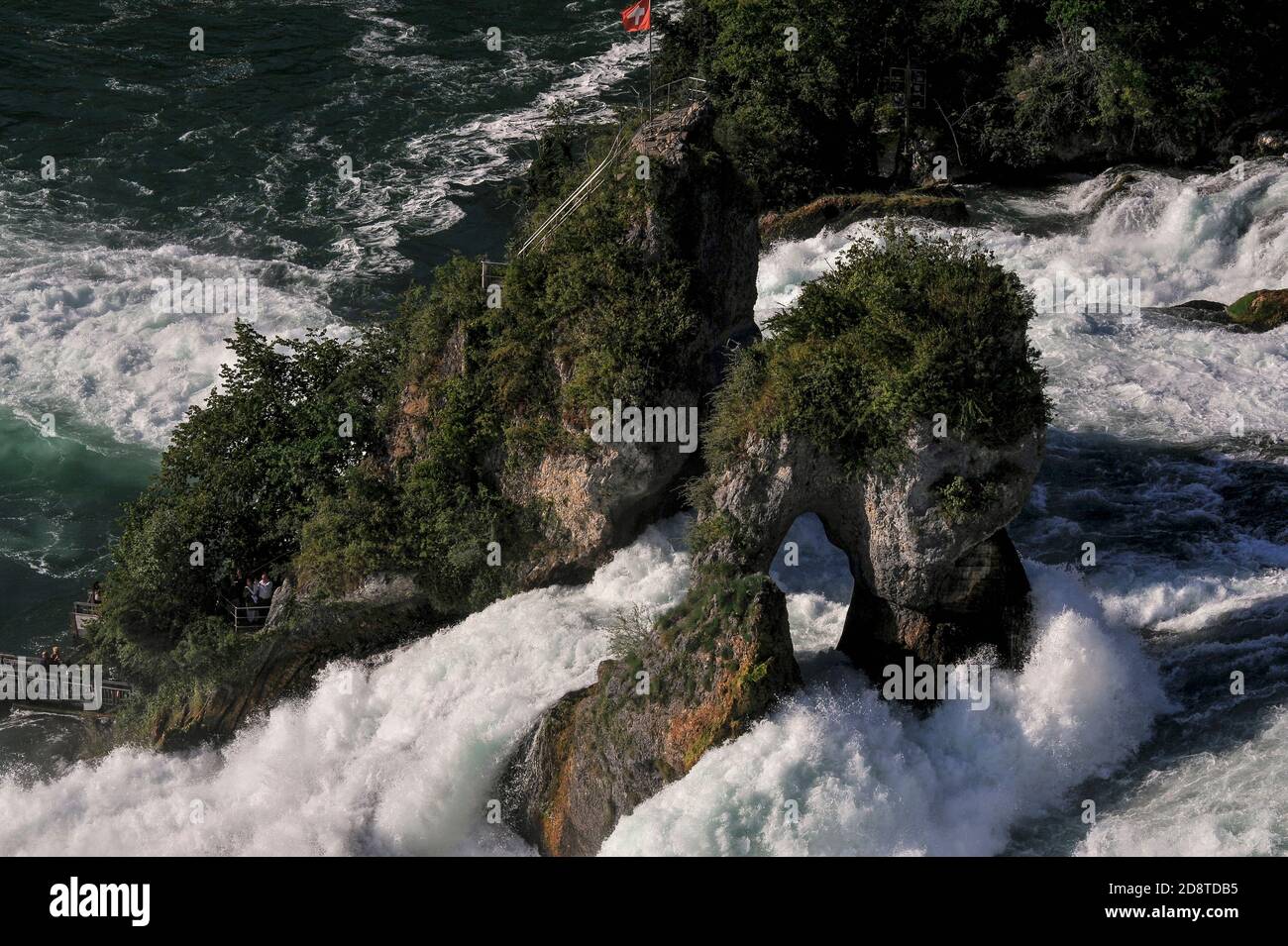 Die Schweizer Flagge fliegt vom Aussichtspunkt auf den Rheinfallfelsen, einem felsigen Felsen, der auf beiden Seiten vom donnernden Rheinfall, dem Rheinfall bei Schaffhausen, der Nordschweiz, dem größten und mächtigsten Wasserfall Europas, passiert wird. Der Rheinfallfelsen, der nur mit dem Boot erreicht wurde, war Teil einer harten Kalksteinklippe, die den ehemaligen Kanal des hoch- oder Oberrheins flankiert. Trotz Erosion hat es Sommerwasserströmungen mit durchschnittlich 600 Kubikmeter (21,000 Kubikfuß) pro Sekunde standgehalten. Der Fluss stürzt etwa 23 m (75 ft), bevor er sich sediger in Richtung Deutschland entwickelt. Stockfoto