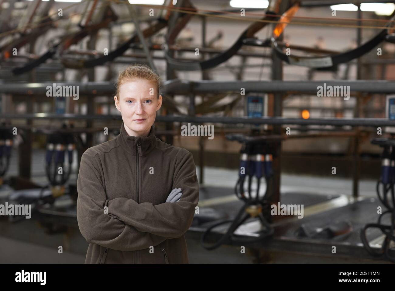 Waist-up-Porträt einer Arbeiterin, die auf die Kamera schaut, während sie mit gekreuzten Armen gegen die industrielle Kuhmelkmaschine in einem modernen Milchviehbetrieb steht, Platz kopieren Stockfoto