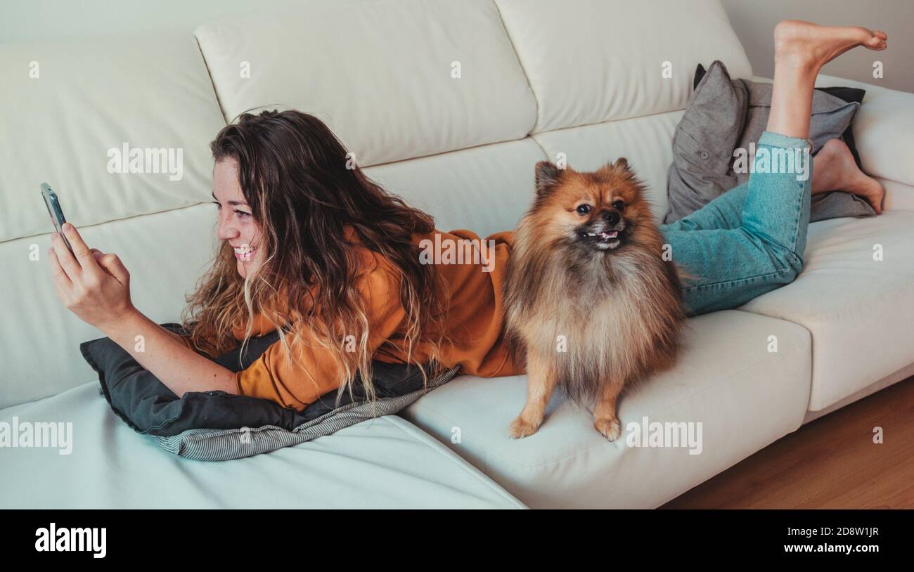 Lächelnde Frau mit dem Telefon, das mit ihrem Hund auf der Couch liegt. Stockfoto