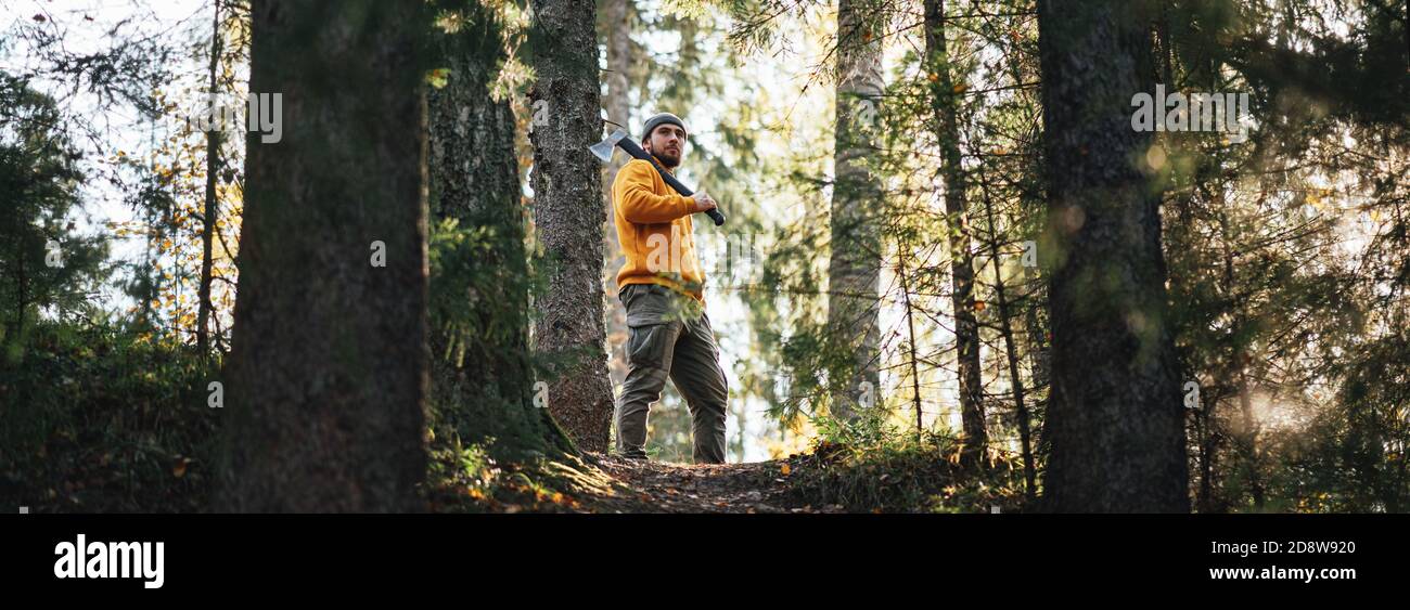 Mann Holzfäller mit Axt im Wald. Starker bärtiger Holzfäller in Arbeitskleidung hält Axt auf seiner Schulter. Breites Bild Stockfoto