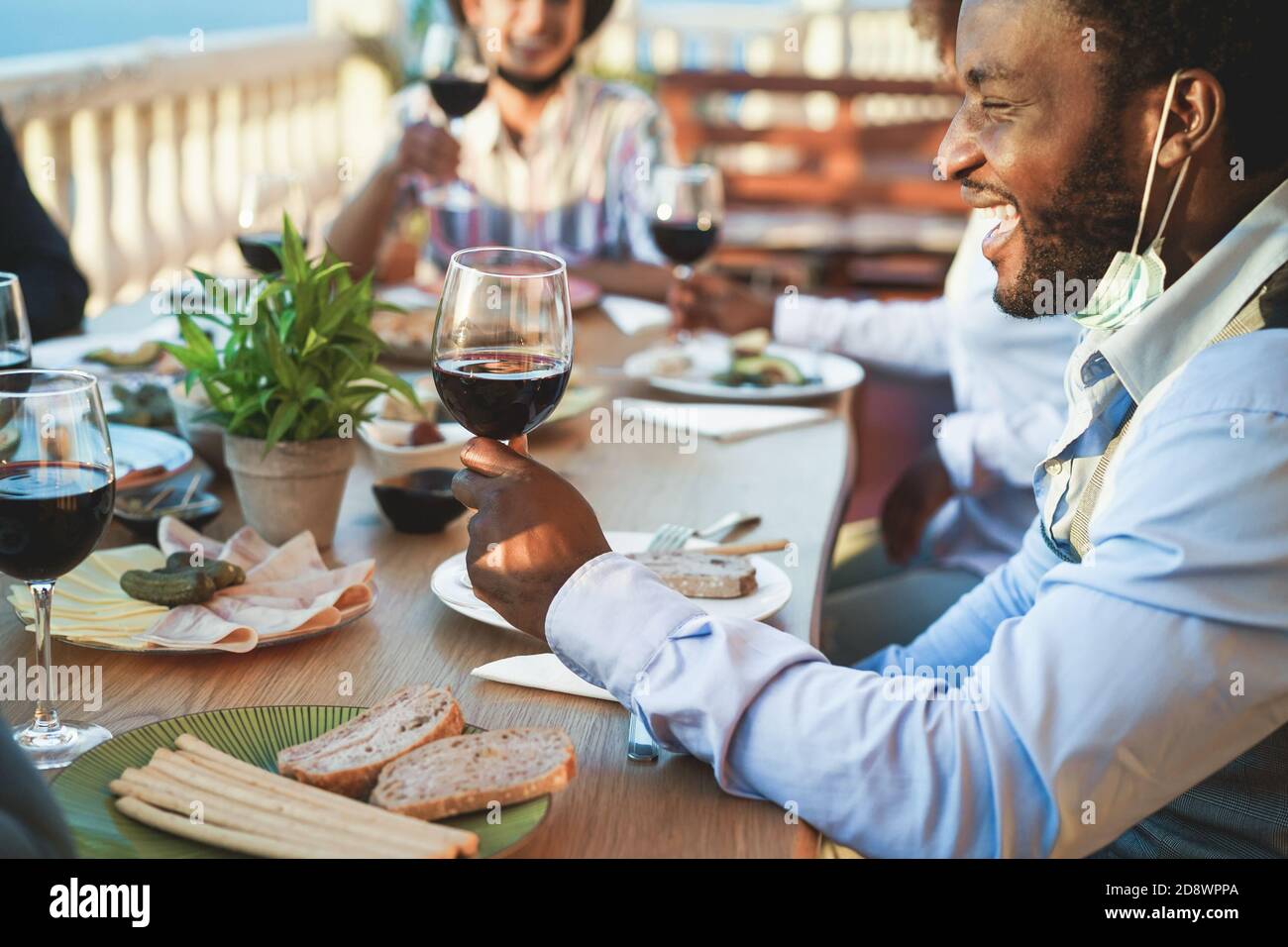 Fröhliche multirassische Freunde essen und trinken Wein, während das Tragen Schutzkleidung Masken - Fokus auf schwarzen Mann Stockfoto