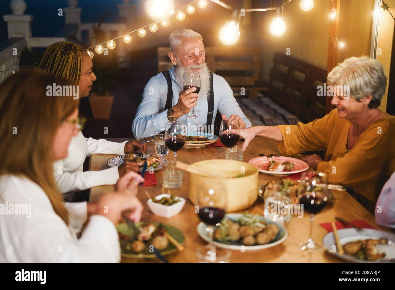 Glückliche ältere Leute, die beim Abendessen auf der Terrasse Spaß hatten - multirassisch Friends Eating on Evening Time - Fokus auf Hipster Mann Gesicht Stockfoto