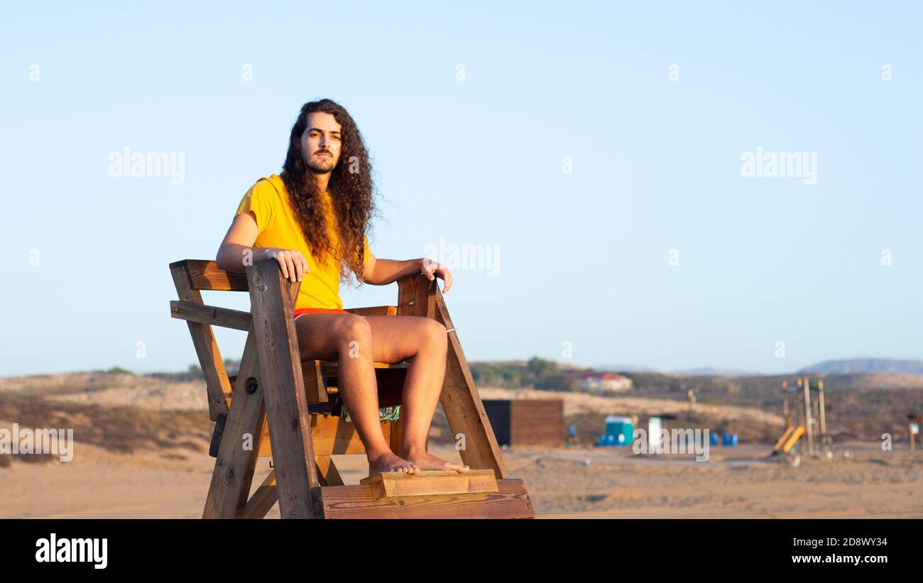 Nahaufnahme eines attraktiven jungen Mannes mit langen Locken Haare sitzen auf einem Rettungsschwimmerstuhl Stockfoto