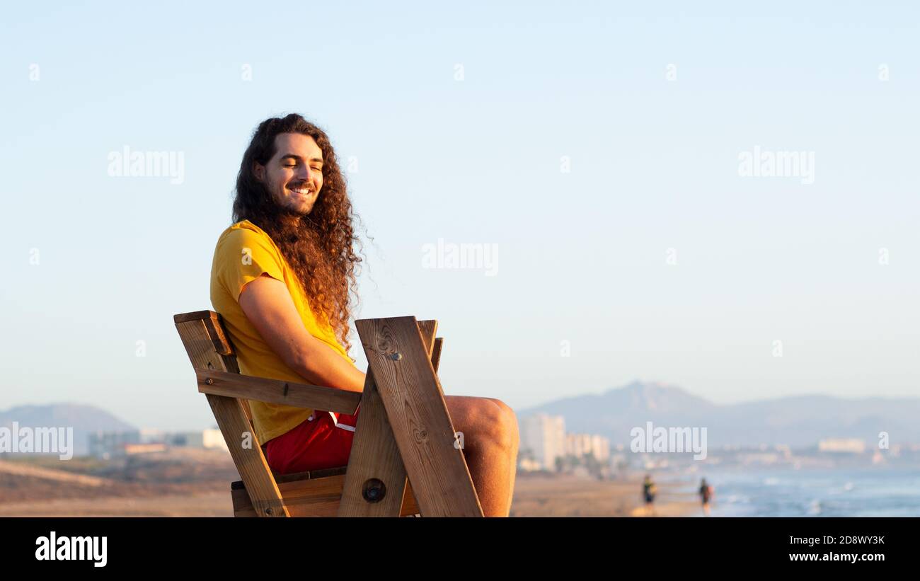 Nahaufnahme eines attraktiven jungen Mannes mit langen Locken Haare sitzen auf einem Rettungsschwimmerstuhl Stockfoto