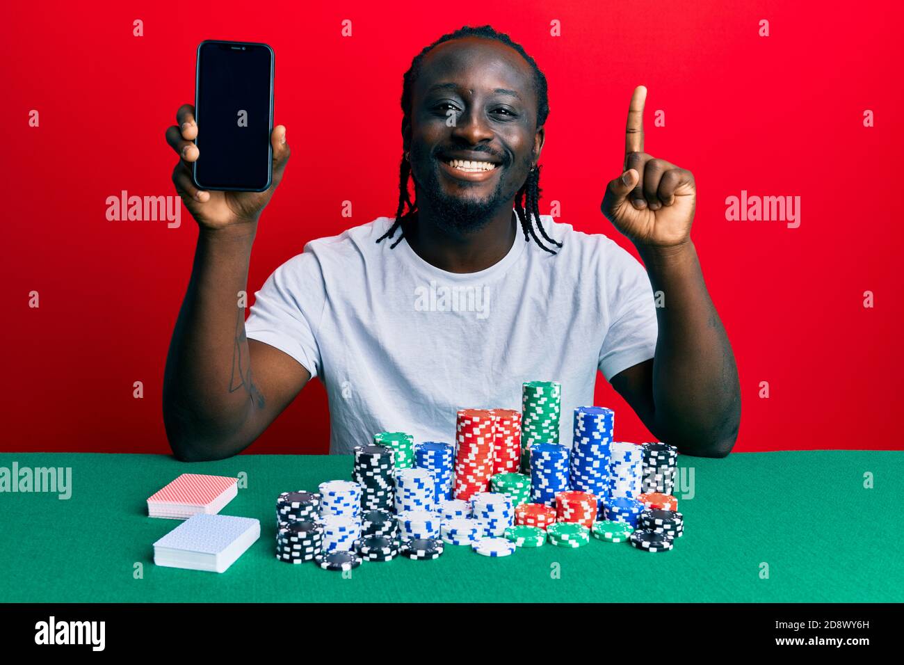 Hübscher junger schwarzer Mann spielt Poker hält Smartphone lächelnd mit einer Idee oder Frage Zeigefinger mit glücklichem Gesicht, Nummer eins Stockfoto