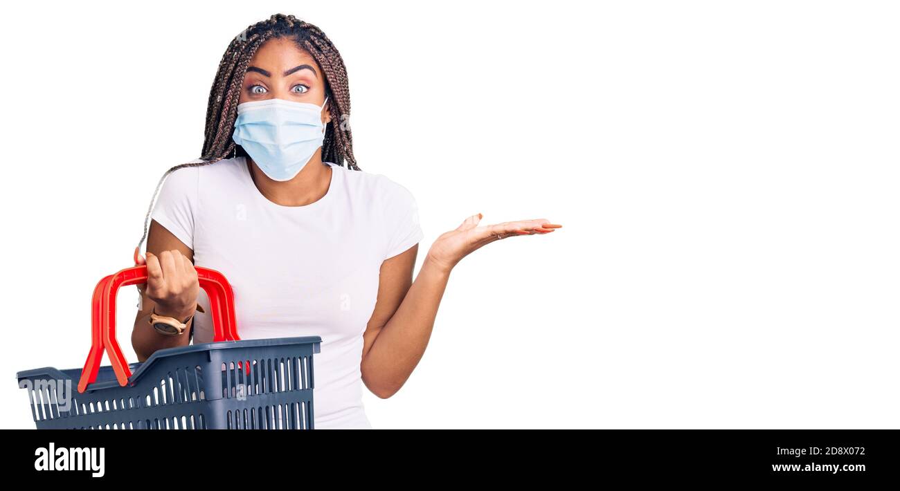 Junge afroamerikanische Frau mit Zöpfen tragen Warenkorb und Medizinische Maske feiert Sieg mit glücklichen Lächeln und Sieger Ausdruck Mit Rais Stockfoto