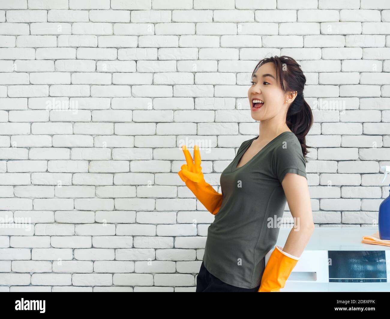 Glücklich fröhlich lächelnde asiatische Frau, Hausfrau trägt orange Gummihandschuhe zeigen zwei Finger, Sieg Zeichen in der Nähe Waschmaschine auf weißen Ziegelwand Stockfoto