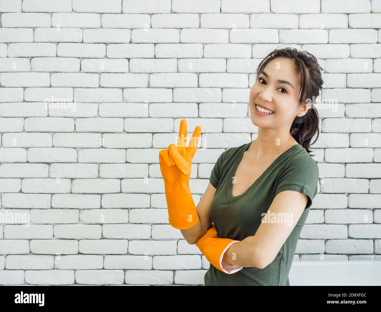Glücklich fröhlich lächelnde asiatische Frau, Hausfrau trägt orange Gummihandschuhe zeigen zwei Finger, Sieg Zeichen in der Nähe Waschmaschine auf weißen Ziegelwand Stockfoto