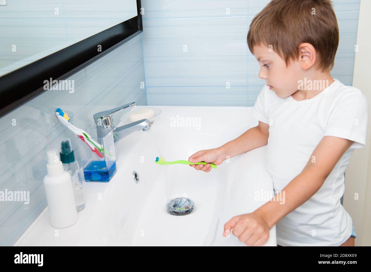 Ein kleines kaukasisches gesundes niedliches Kind in weißen Hemdwäsche Seine grüne Zahnbürste unter frischem Wasser lehnte sich auf das Waschbecken Comfort blaues Badezimmer bei Stockfoto