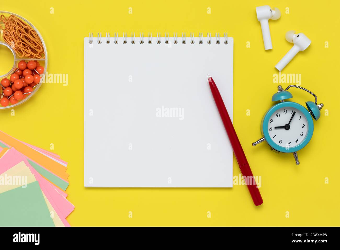 Notizbuch und Stift auf gelbem Hintergrund. Notizblock, weiße Kopfhörer, Wecker, Clips und bunte Aufkleber am Arbeitsplatz. Vorlage mit Kopierbereich, Stockfoto